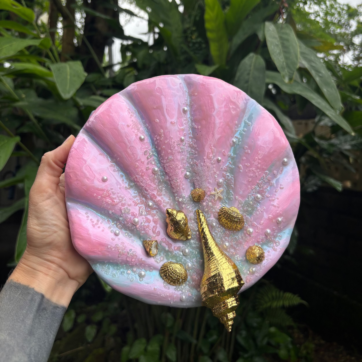 Round, pink and Gold Textured Art Piece held in front of green plants